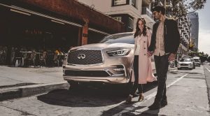 A woman and a man standing in the street to the side of the front of a parked champagne colored 2020 INFINITI QX80. | INFINITI dealer in Ellisville, MO